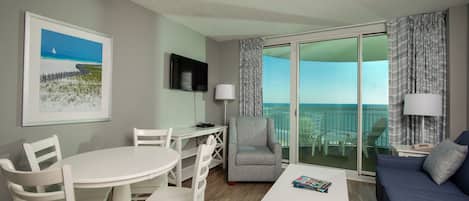 A modern hotel room with a balcony, white furniture, a flat-screen TV, and a painting of a beach scene.