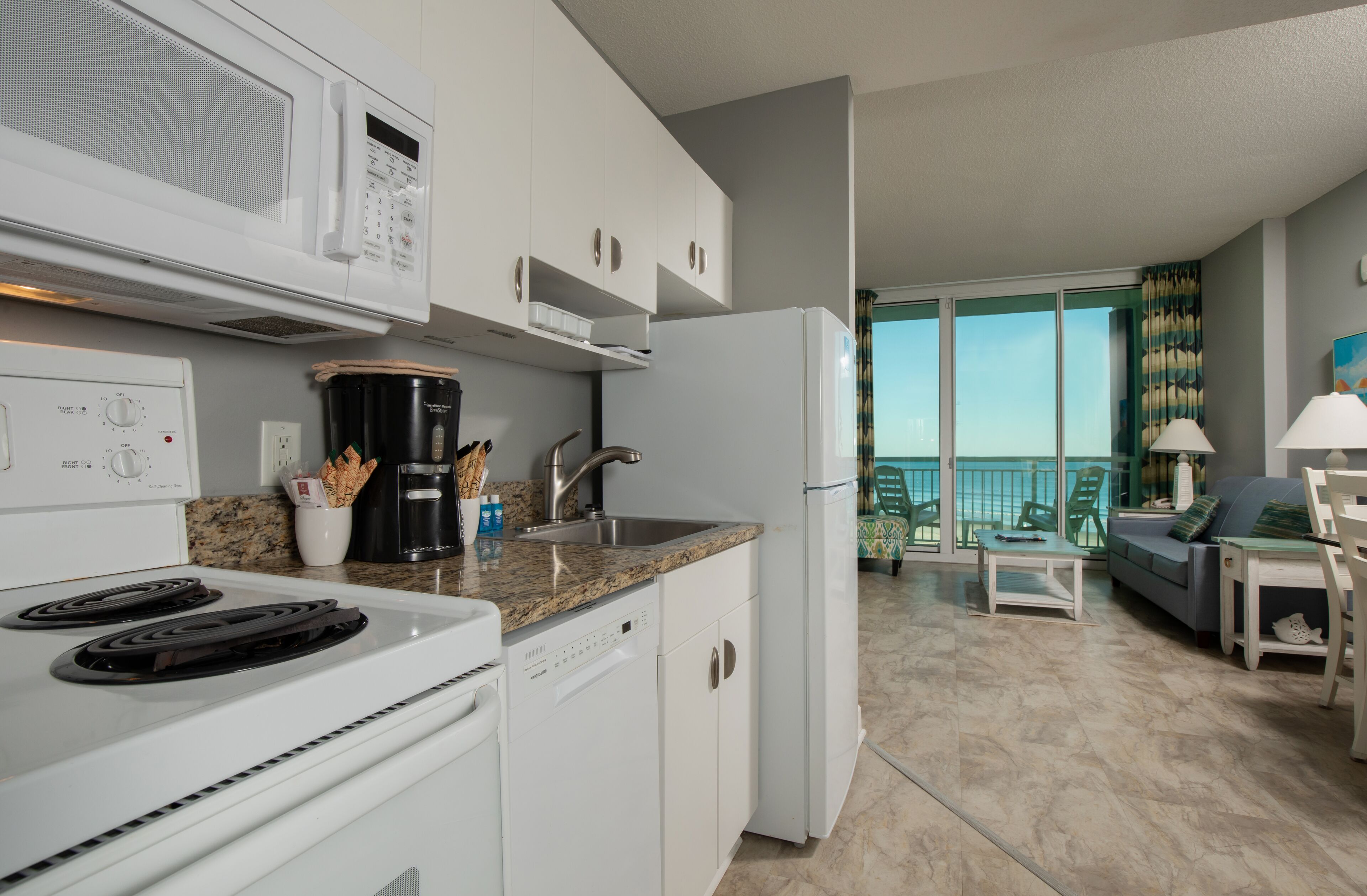 A modern kitchen with white appliances, a granite countertop, and a view of the balcony.