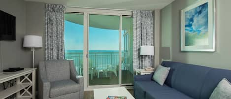 Una sala de estar moderna con un sillón gris, un sofá azul y una mesa de centro blanca. Hay una puerta de cristal que da a un balcón con vistas al cielo y un cuadro en la pared.