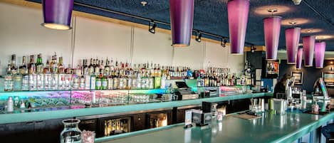 A bar with a long counter, various bottles, and purple hanging lights.