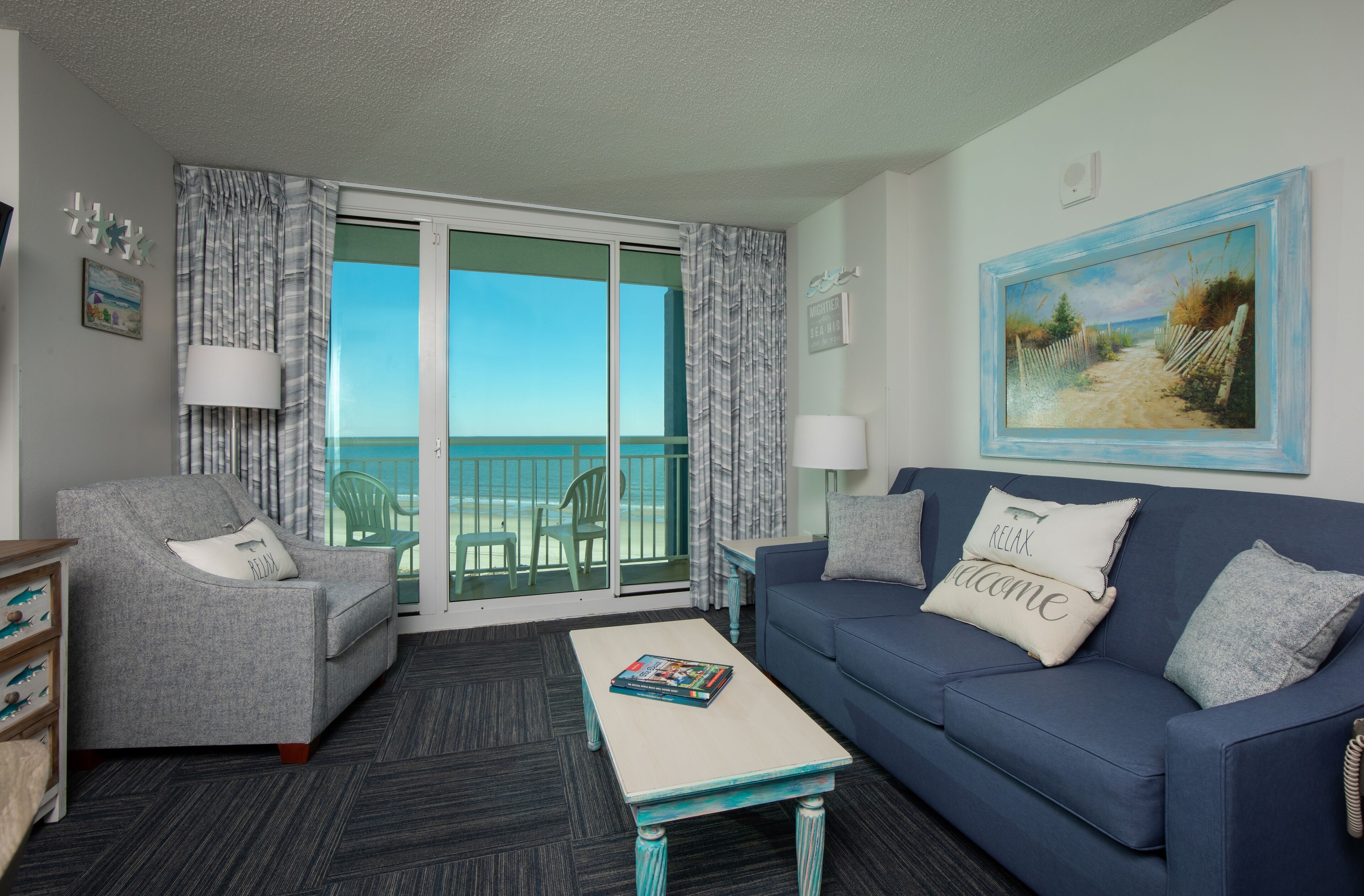 A room with a grey sofa, a blue sofa, a coffee table, and a view of the ocean through a sliding glass door.