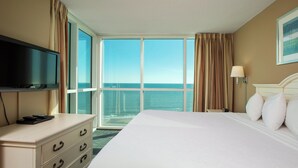 A hotel room with a large bed, a television on a dresser, a nightstand with a lamp, and a view of the ocean through large windows. - Avista Resort (North Myrtle Beach)