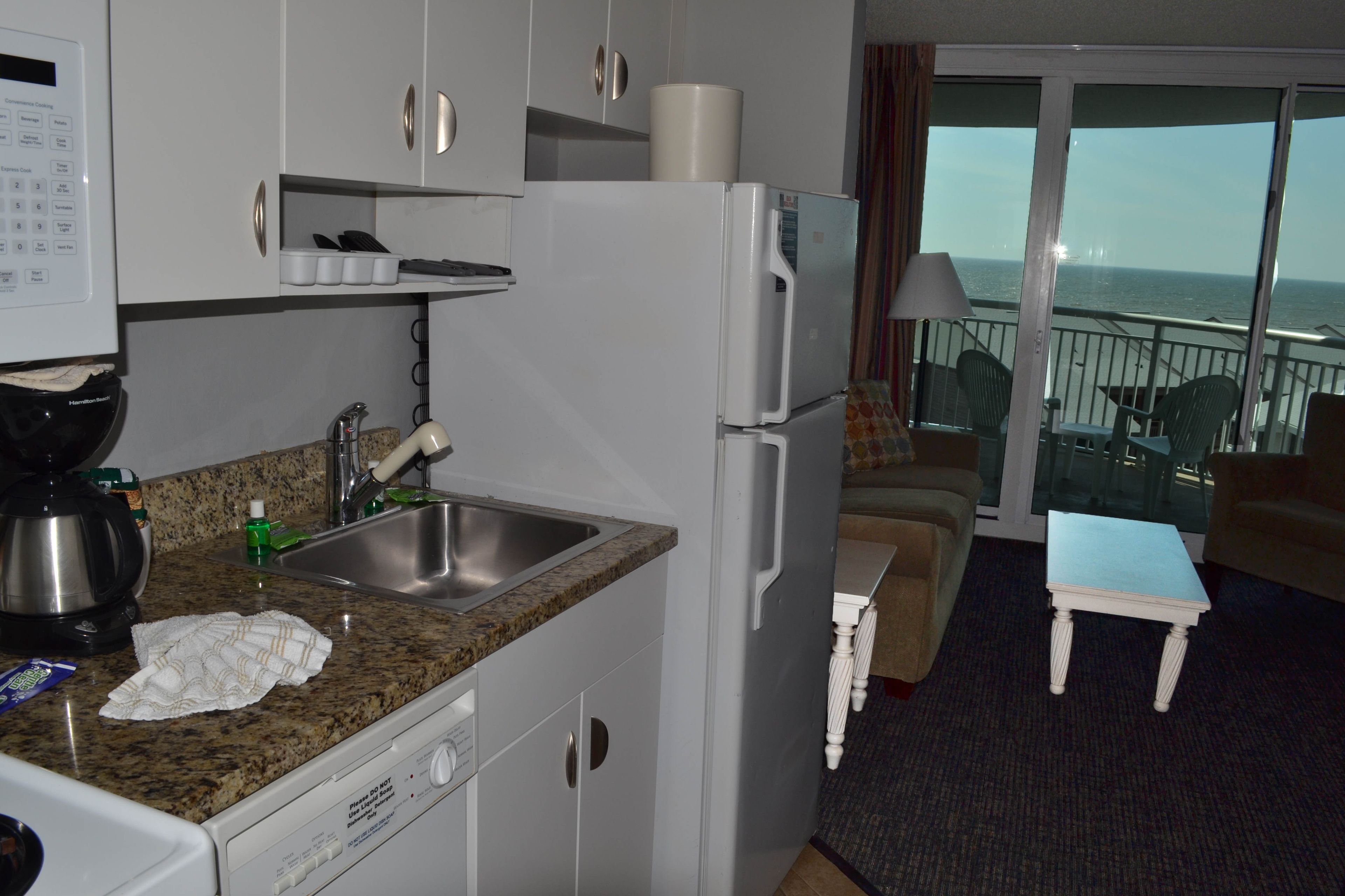 Eine kompakte Küche mit Granit Arbeitsplatte, Edelstahlspüle und Kühlschrank. Ein kleiner Essbereich mit Tisch und Stühlen ist zu sehen, ebenso ein Balkon mit Blick aufs Meer.