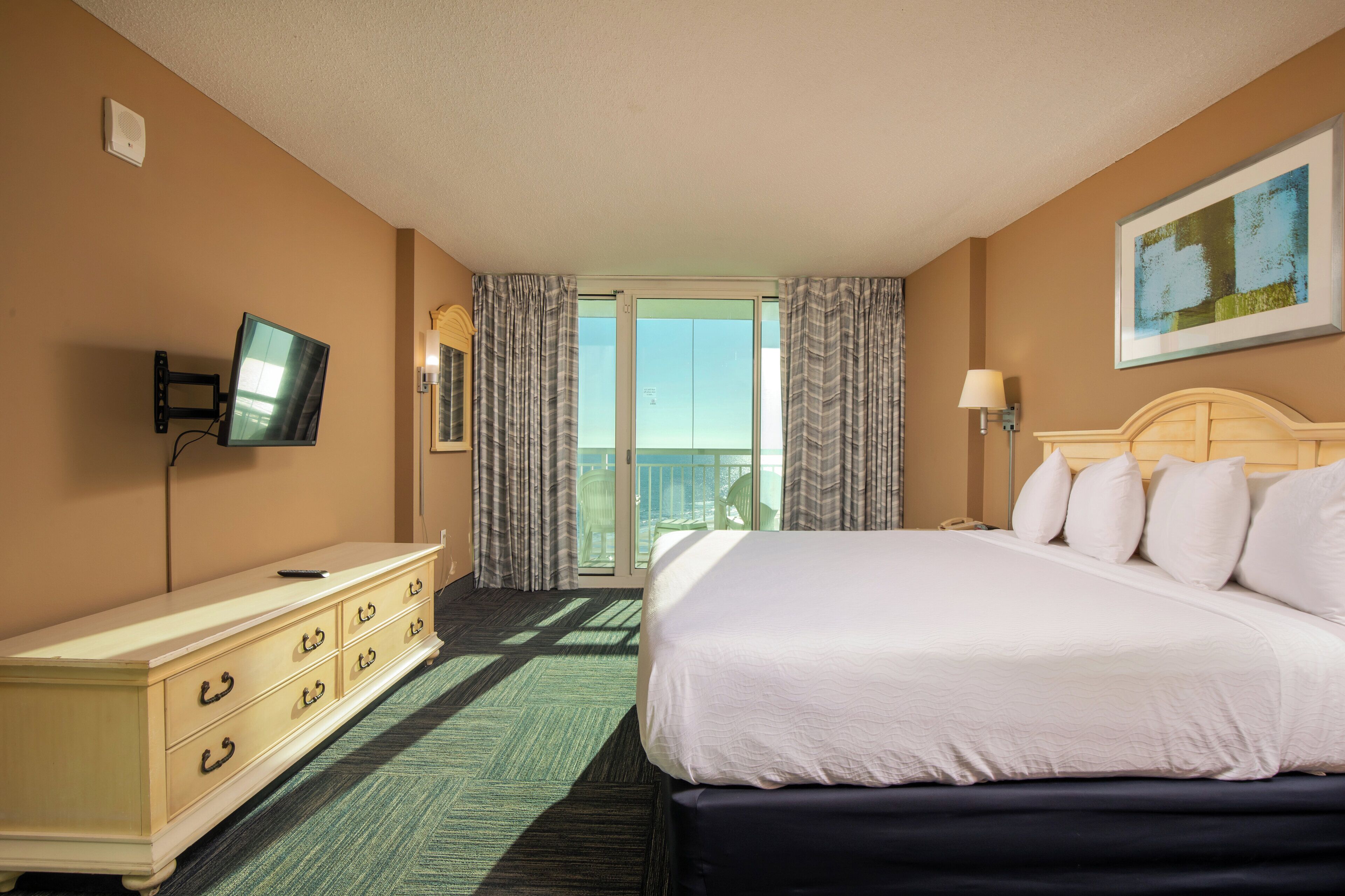 A hotel room with a bed, a dresser, a television, a balcony with curtains, and a painting on the wall.