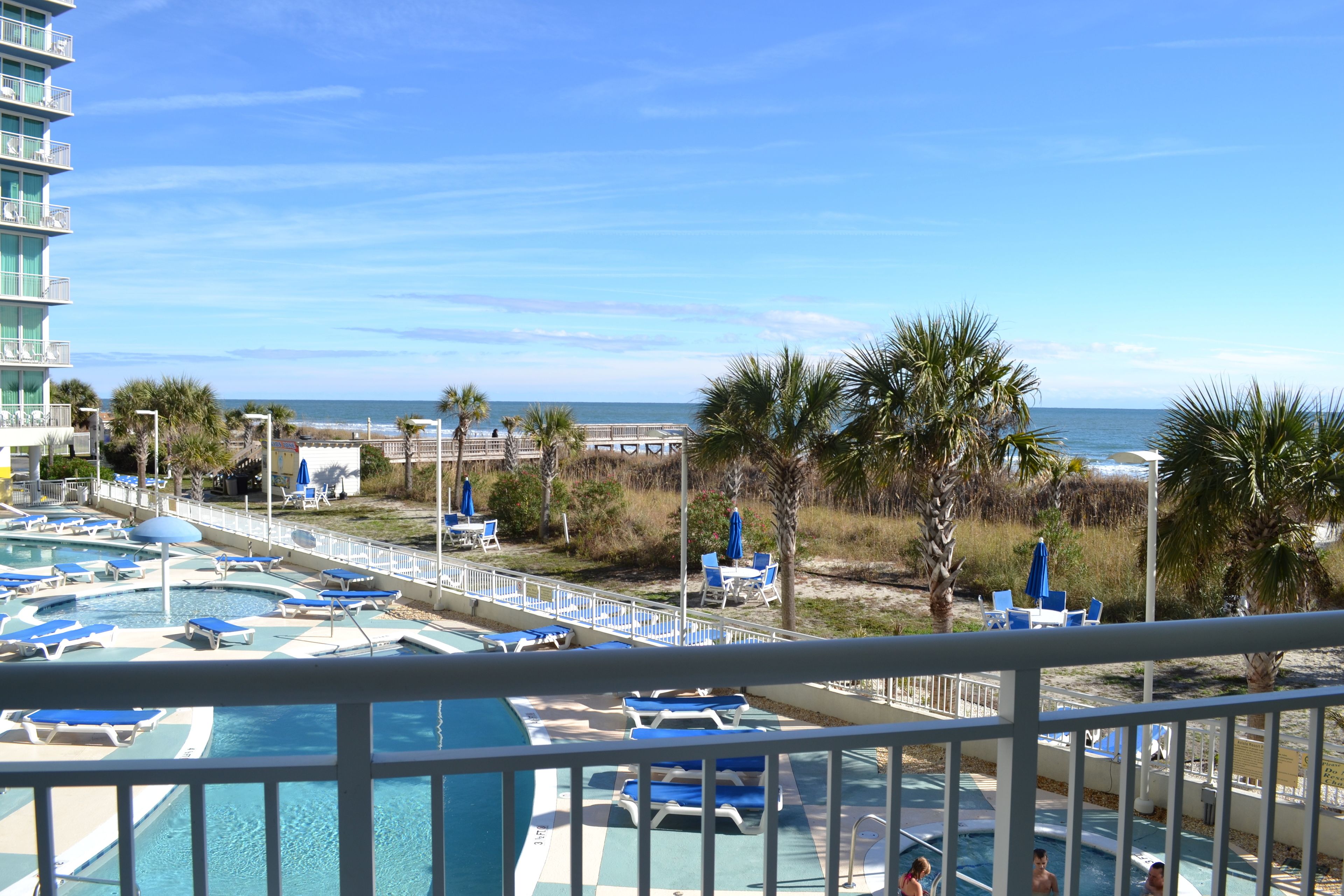 A pool area with multiple pools and lounge chairs, a beach in the background, and a building with balconies.