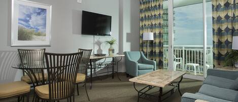 A living room with a glass table, chairs, a sofa, a flat-screen TV, and a balcony with patterned curtains.