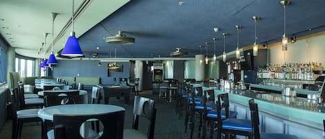 Un espace bar offrant différentes options de sièges, notamment des tables rondes et des tabourets de bar, ainsi qu’un comptoir bien garni de bouteilles variées.