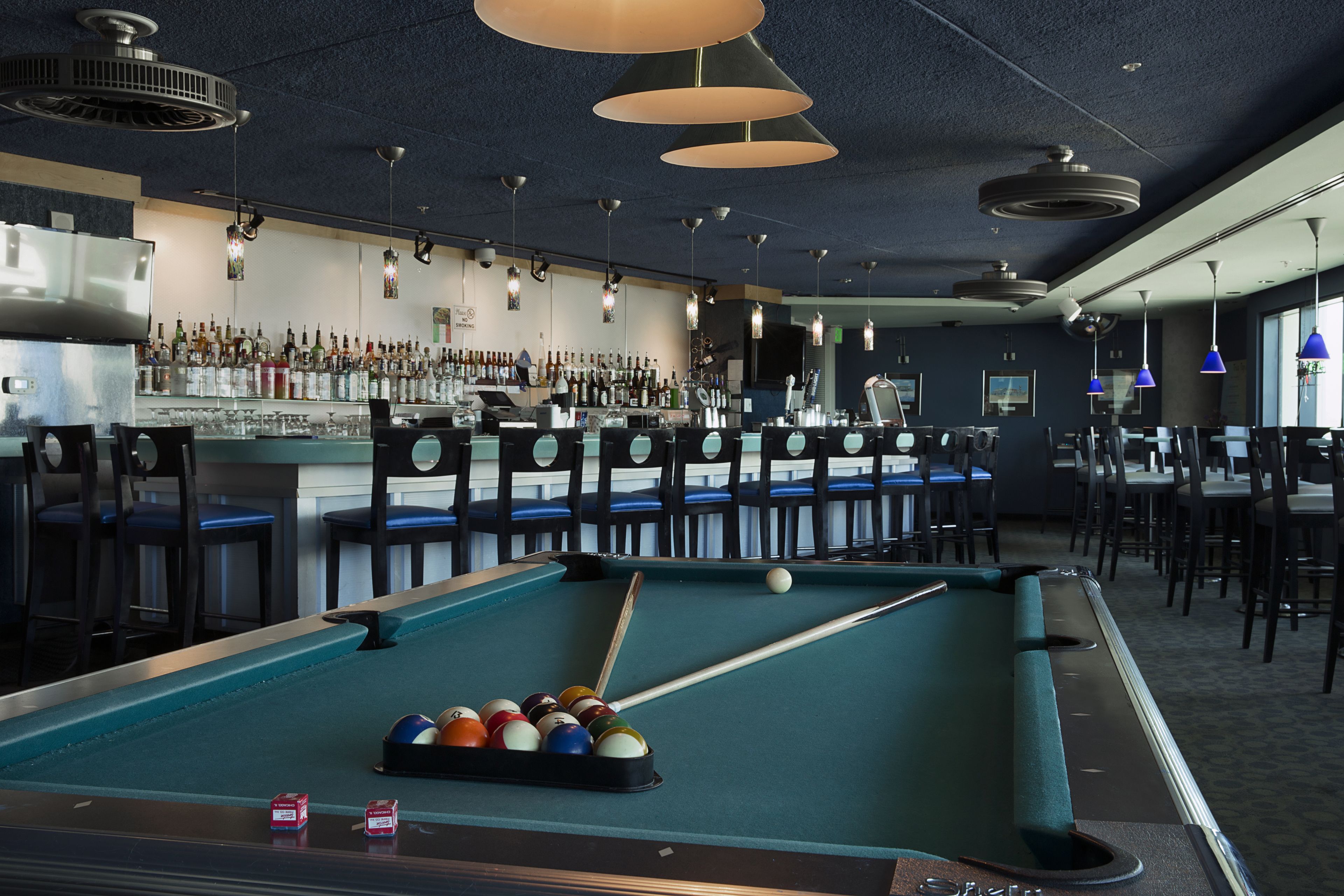 Une table de billard avec des billes et des queues, un bar avec des bouteilles et différents types de sièges.