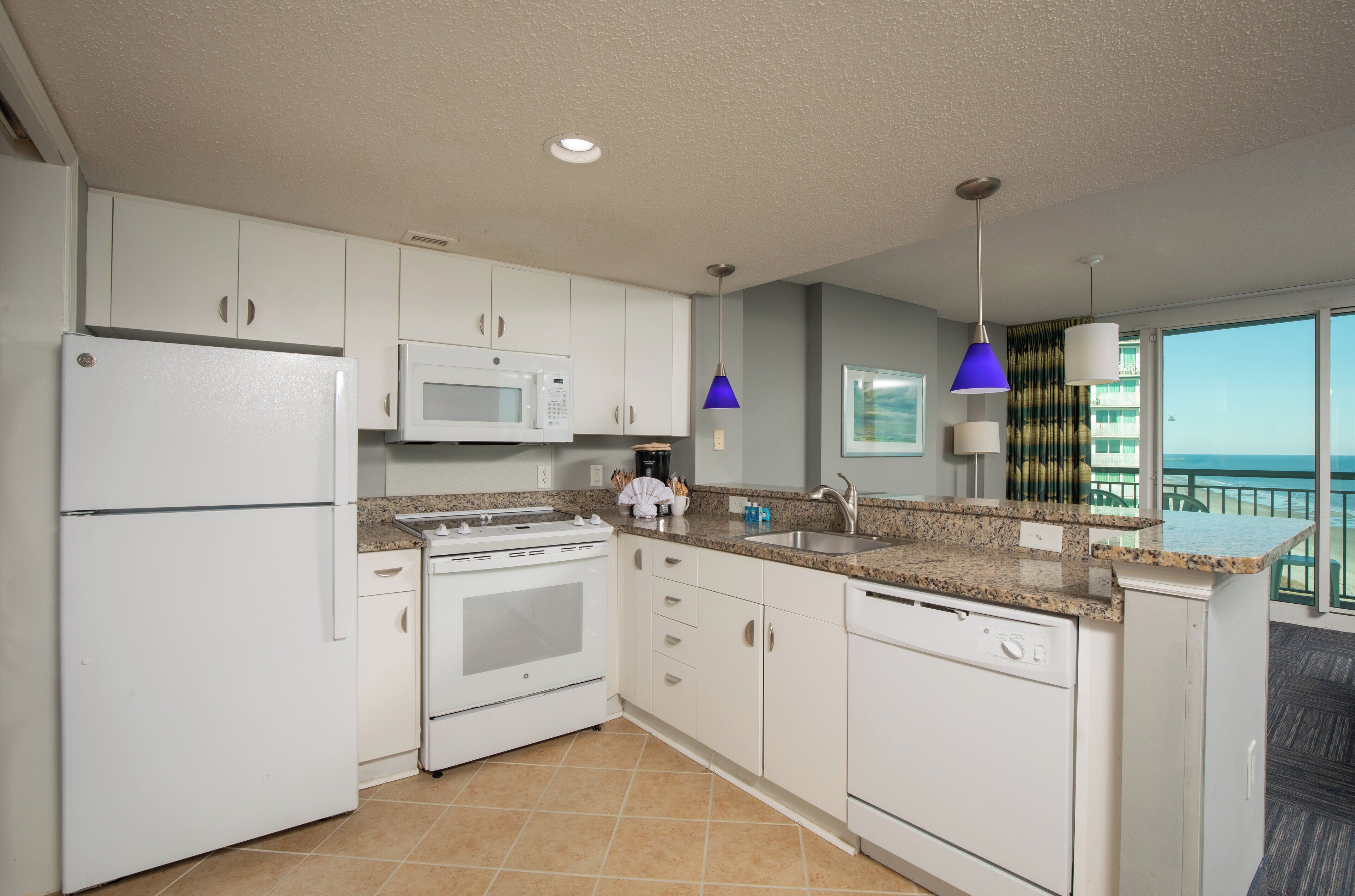 A modern kitchen with white cabinets, a microwave, a stove, and a dishwasher. There is a large window offering a view of the ocean.