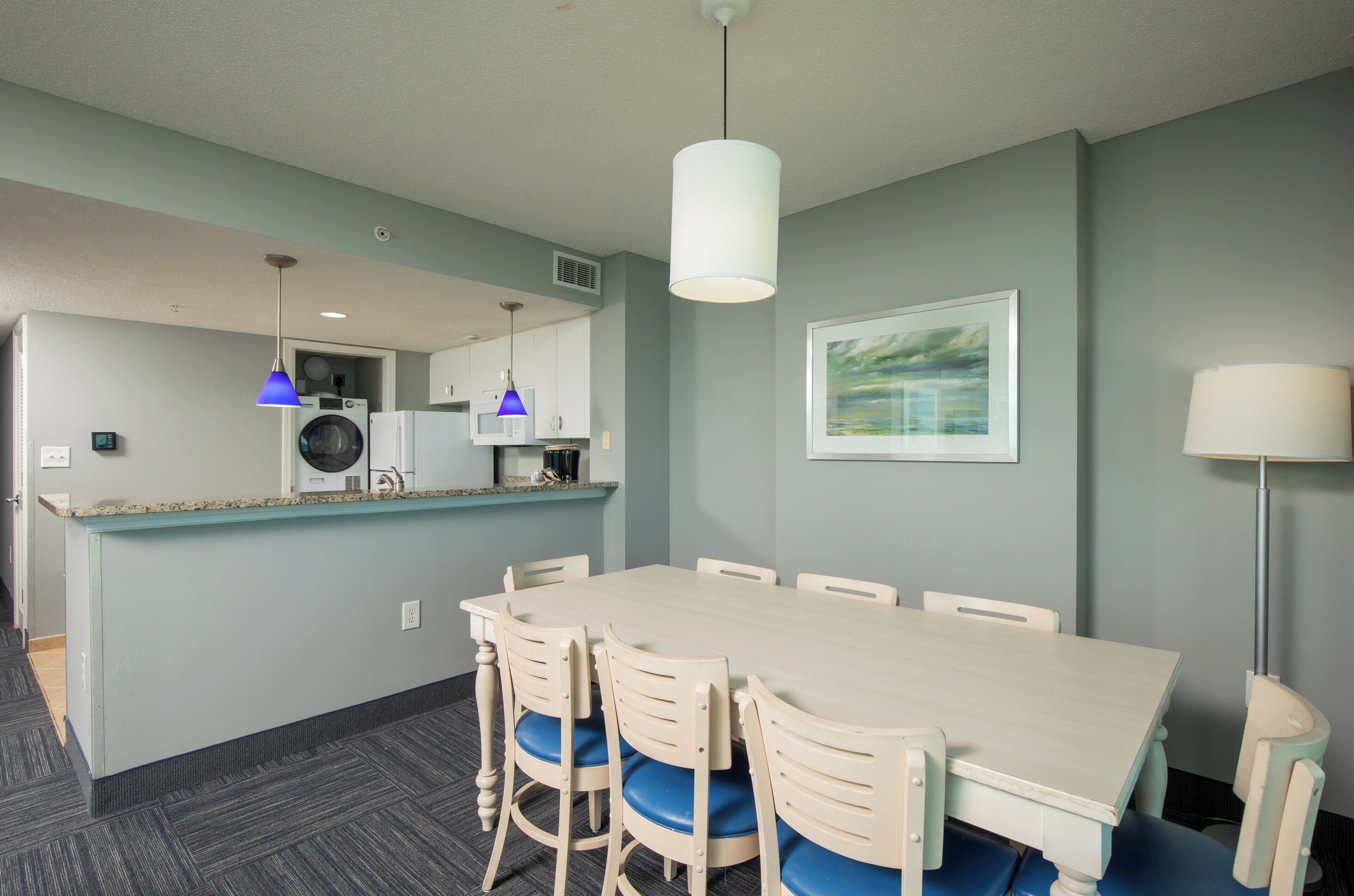 A modern kitchen with a built-in washer and dryer, a dining area with a large table and chairs, and a pendant light above the table.