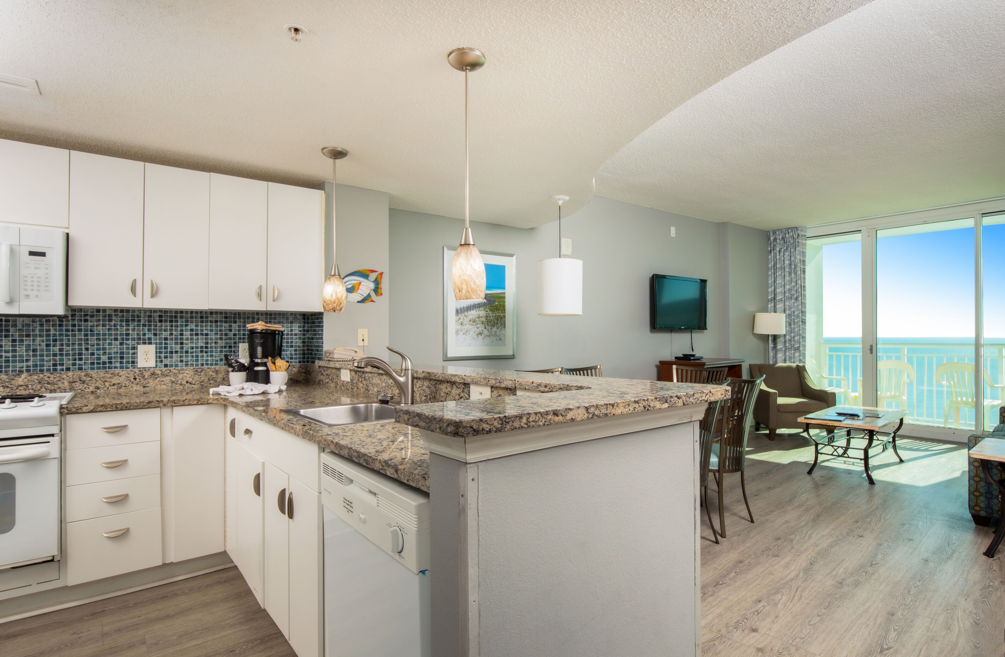 A modern kitchen with granite countertops, white cabinets, and a built-in dishwasher. There is a dining area with a table and chairs, and a large window offering a view of the ocean.