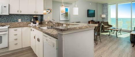 A modern kitchen with granite countertops, white cabinets, and a built-in dishwasher. There is a dining area with a table and chairs, and a large window offering a view of the ocean.