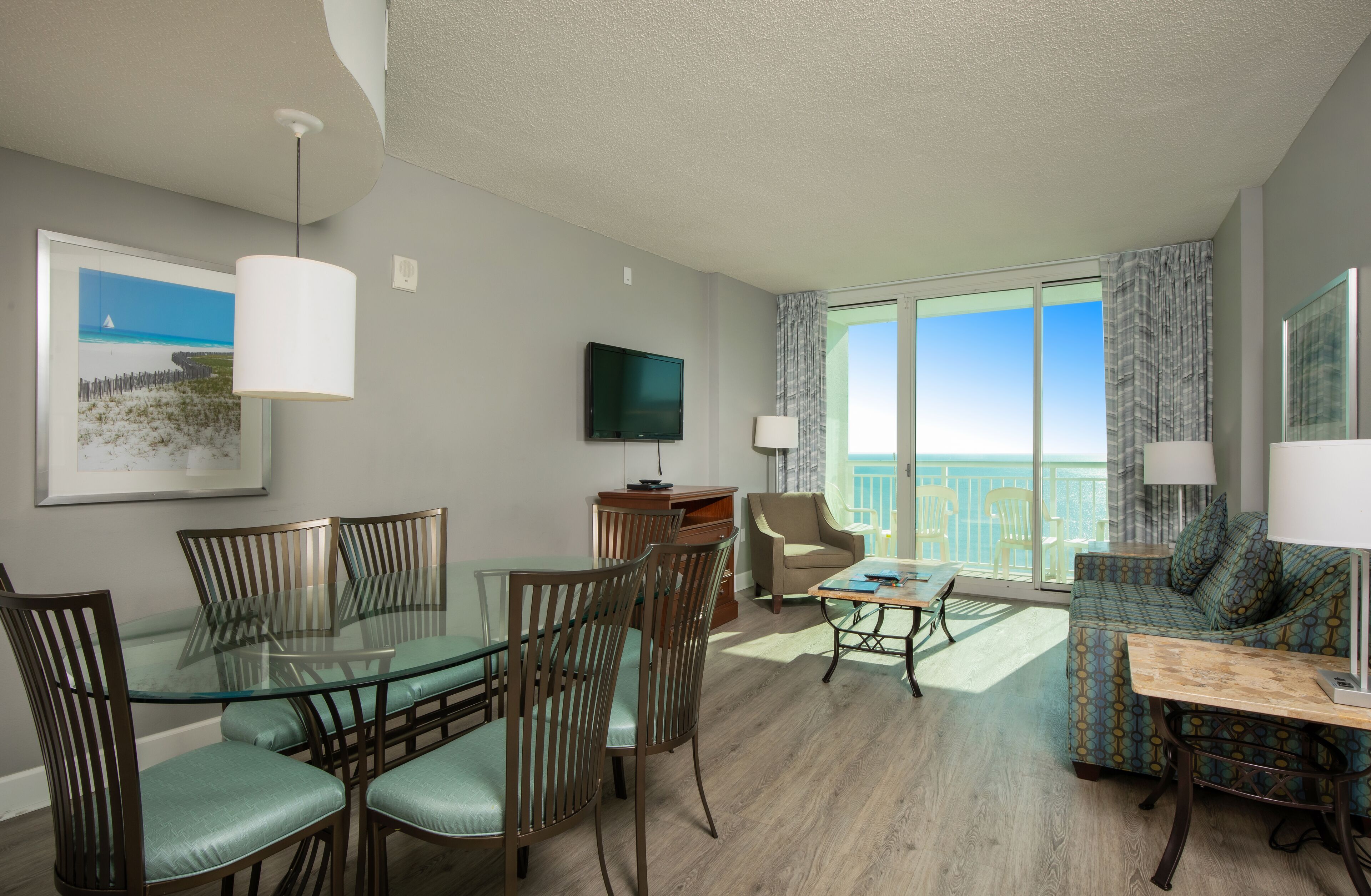 A modern hotel room with a glass dining table, comfortable seating, a flat-screen TV, and a balcony with a view of the beach.