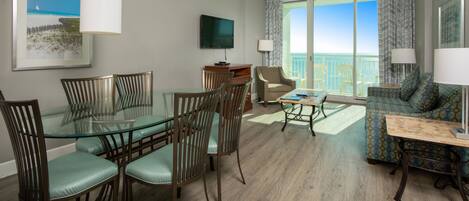 A modern hotel room with a glass dining table, comfortable seating, a flat-screen TV, and a balcony with a view of the beach.