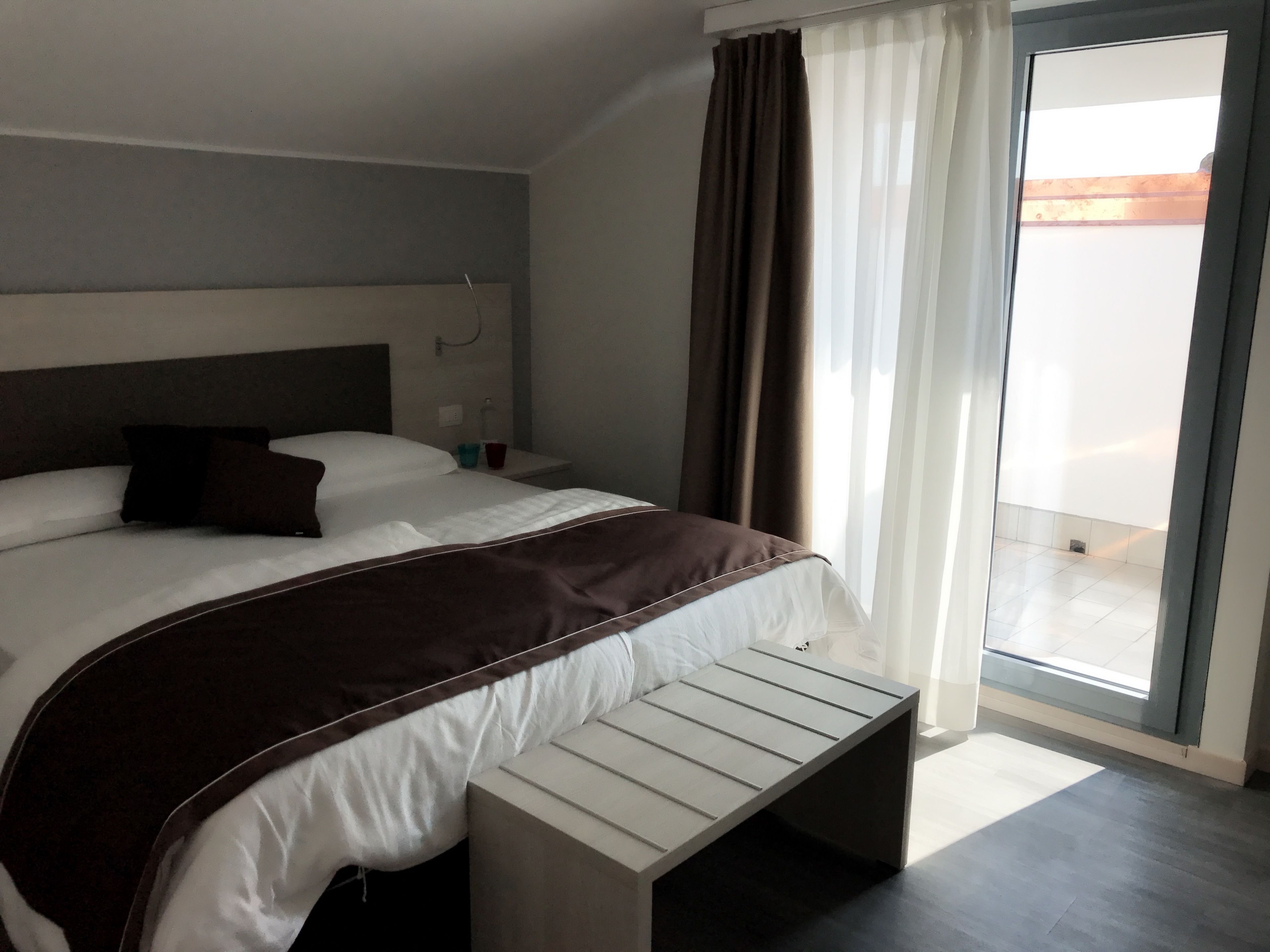A modern hotel room with a large bed, a bedside table, a bench, and a view of the outside through a sliding glass door.