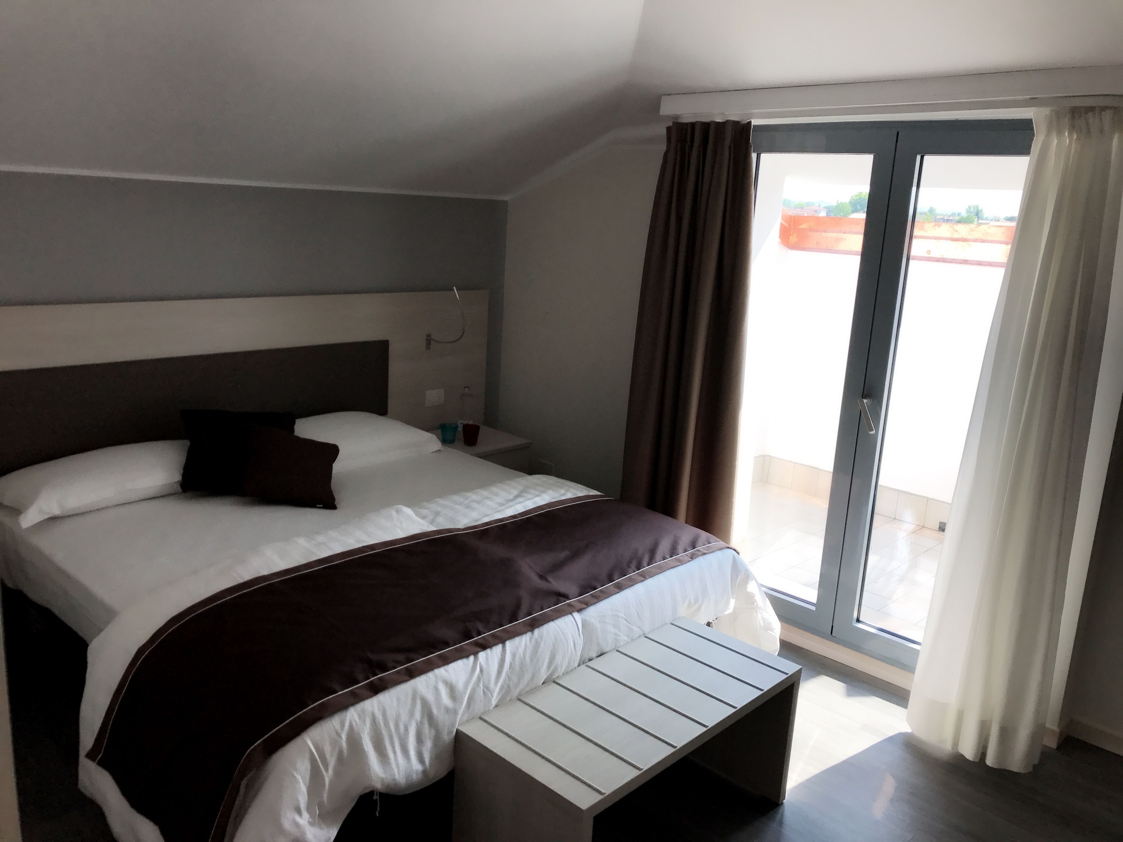 A modern hotel room with a large bed, a bedside table, a small table, and a view of the outside through a sliding glass door.