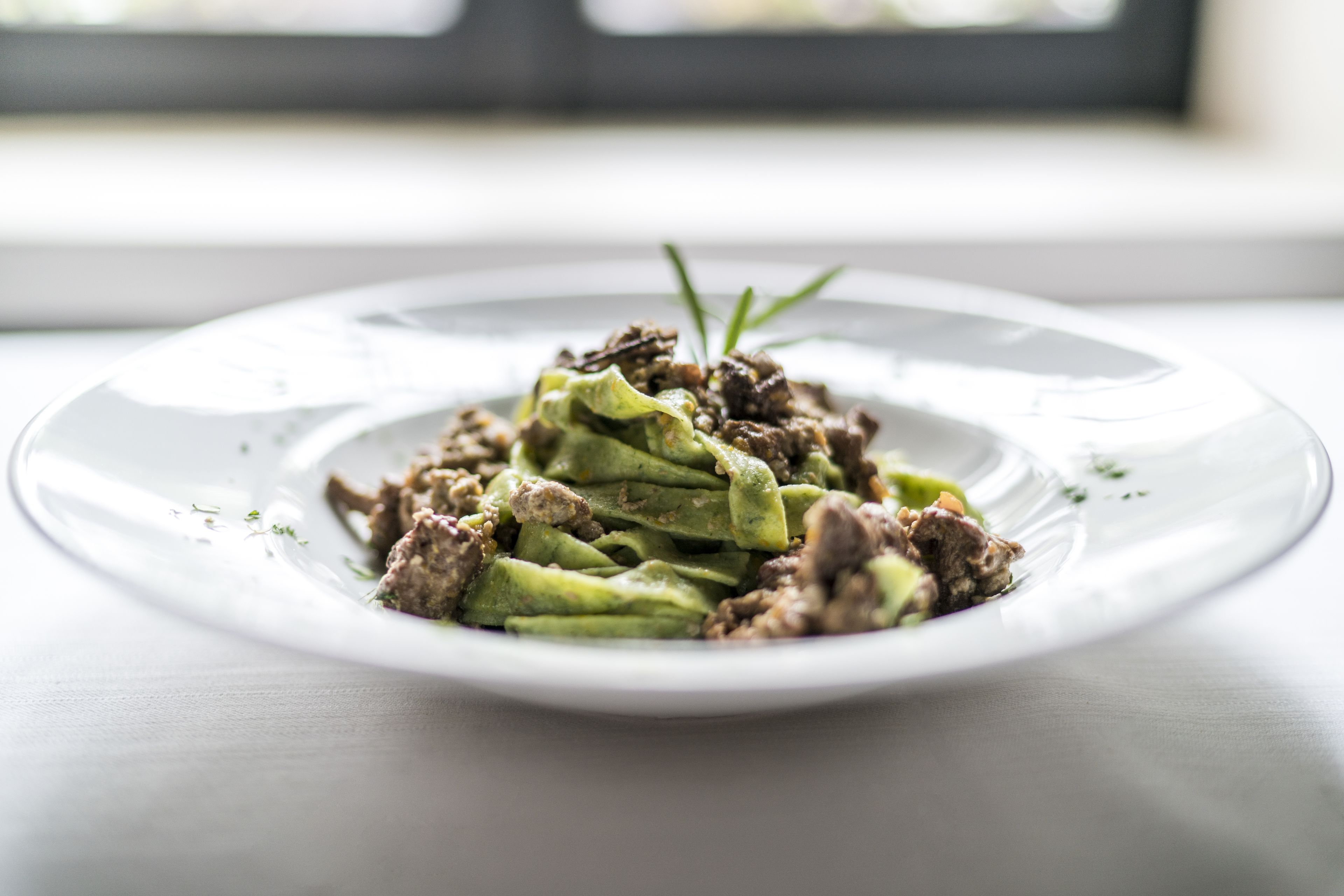 A white plate with a dish of meat and green vegetables, garnished with a sprig of herb, on a light-colored table.