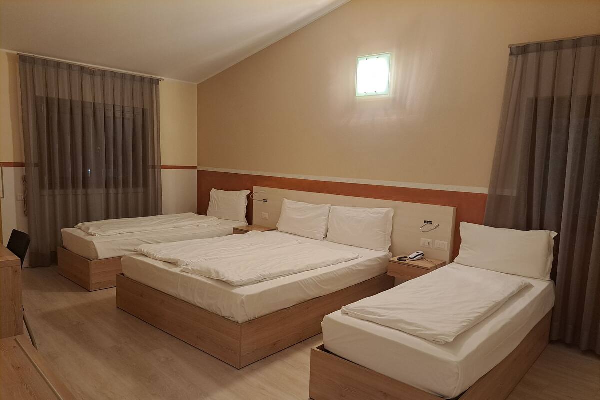 Ein Hotelzimmer mit drei Einzelbetten, einem Holznachtkästchen, einem Schreibtisch und einem großen Fenster mit Vorhängen.