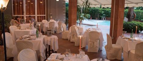 A restaurant with tables set for dining, a pool in the background, and a covered outdoor area.