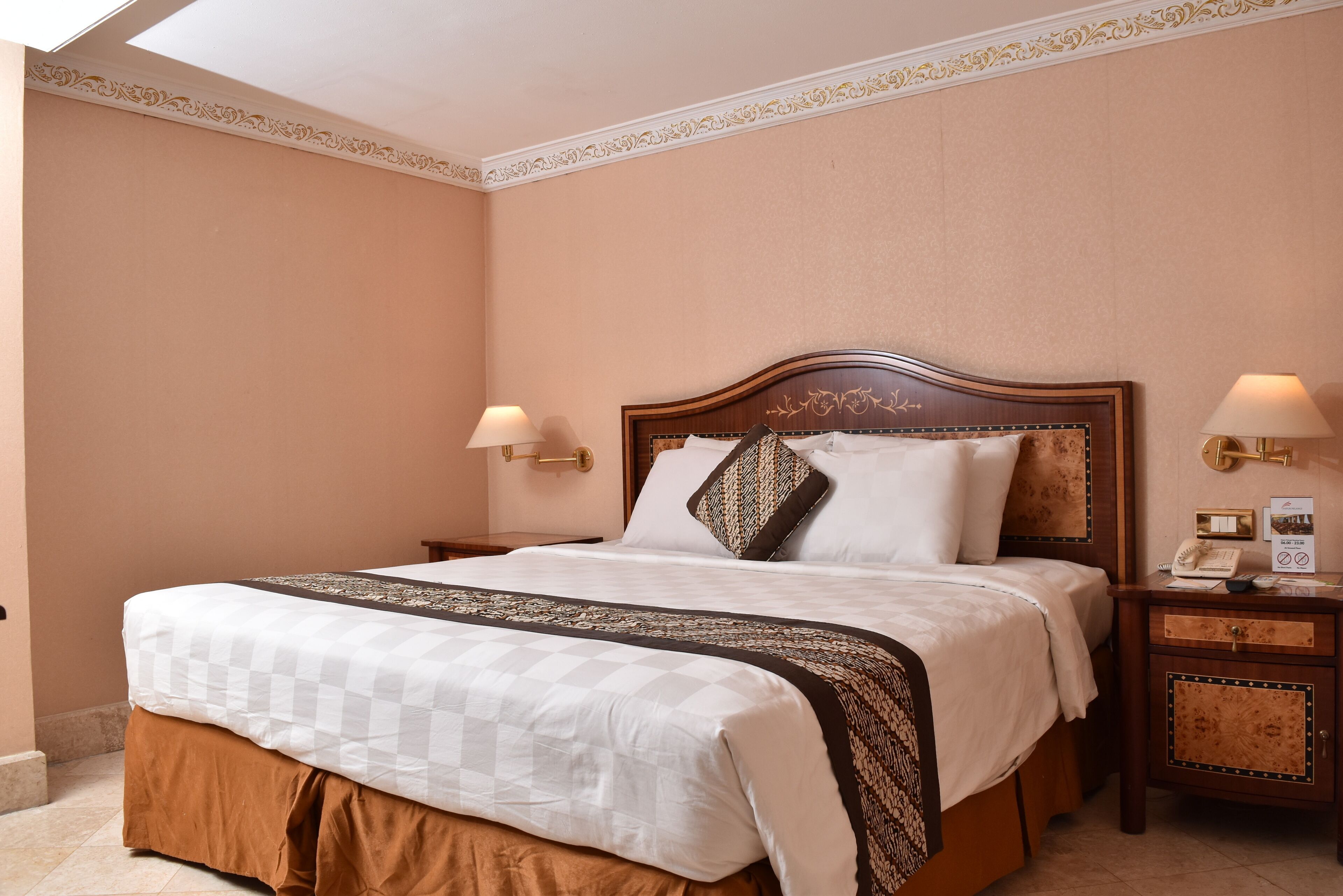 A neatly made bed with a checkered bedspread, a brown skirt, and a decorative headboard. There are two bedside tables with lamps and a small nightstand with a telephone and a sign.