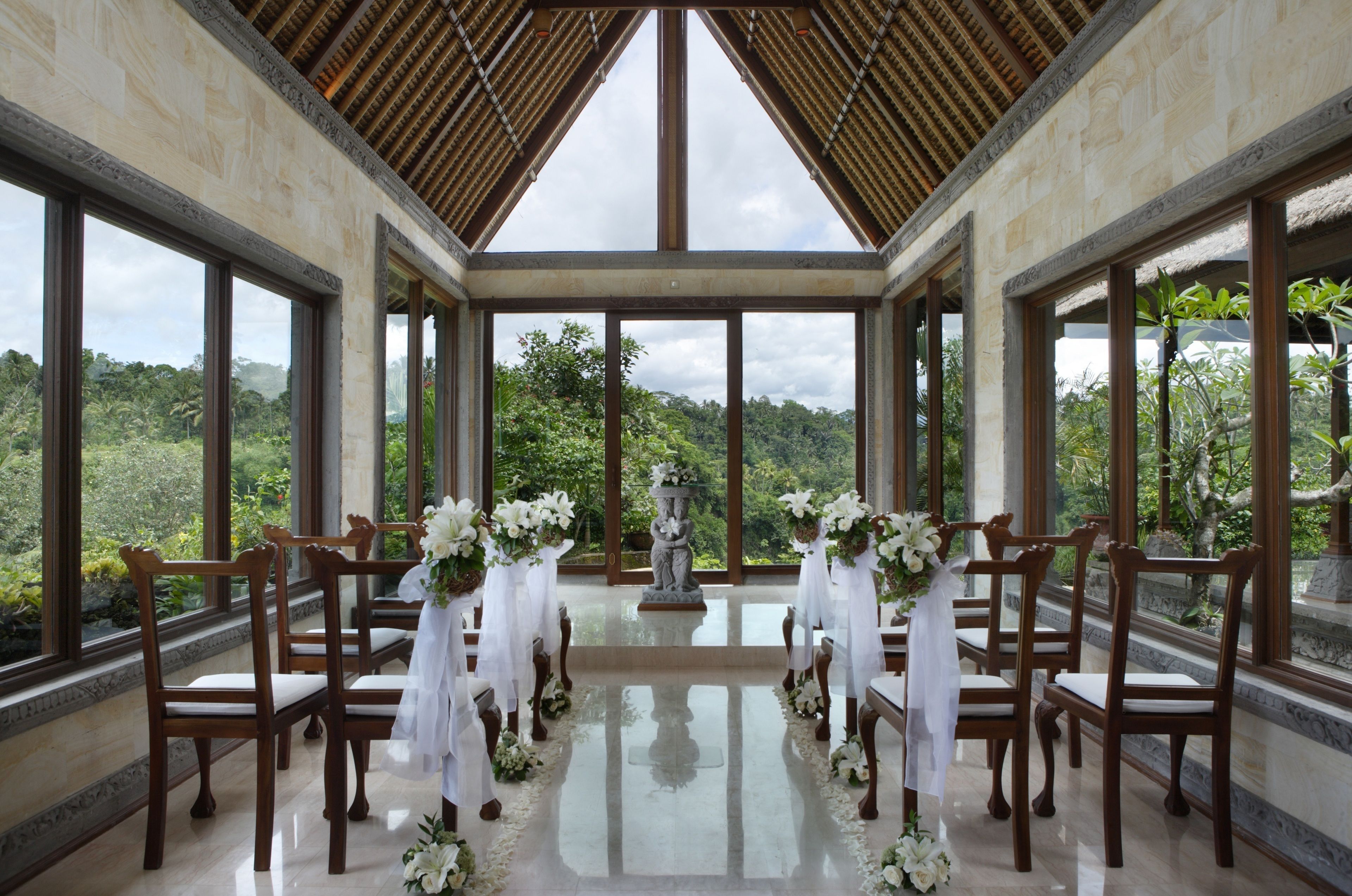 A wedding ceremony setup with wooden chairs and a central aisle decorated with white flowers, large windows, and a view of a lush landscape.