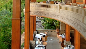 An outdoor restaurant with wooden columns and tables set for dining, overlooking a lush garden. - Royal Pita Maha (Ubud)