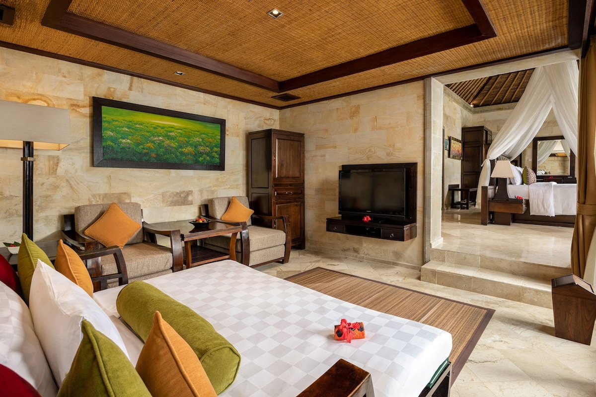 A hotel room with a large bed, a TV, a painting, a desk, and a separate bedroom area with a bed and a nightstand.