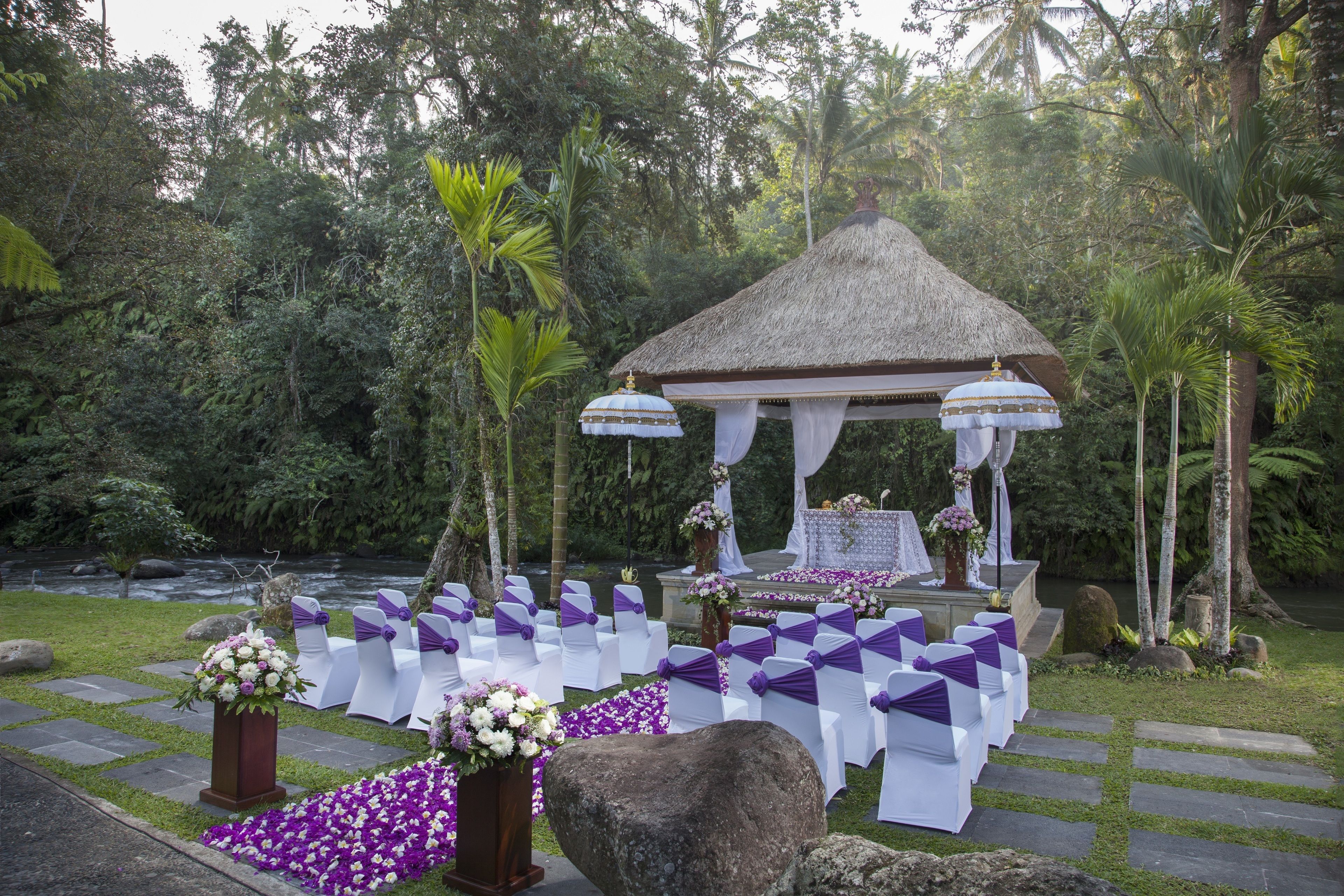 Decoración para una boda al aire libre con un pabellón de paja, sillas blancas con cintas moradas y arreglos florales.