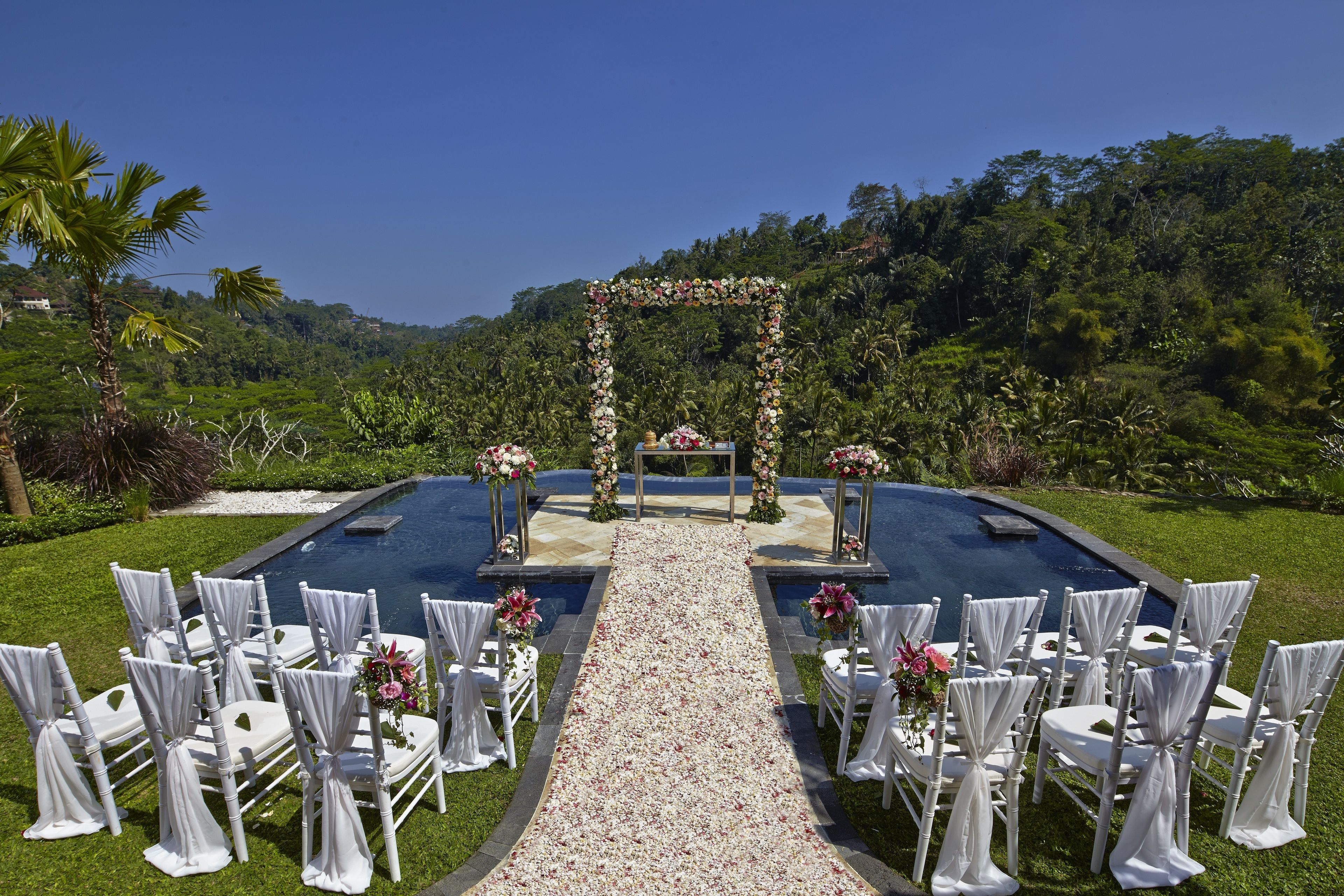 Ceremonia de boda al aire libre con un arco floral y sillas blancas dispuestas sobre un camino de grava que lleva a una piscina.