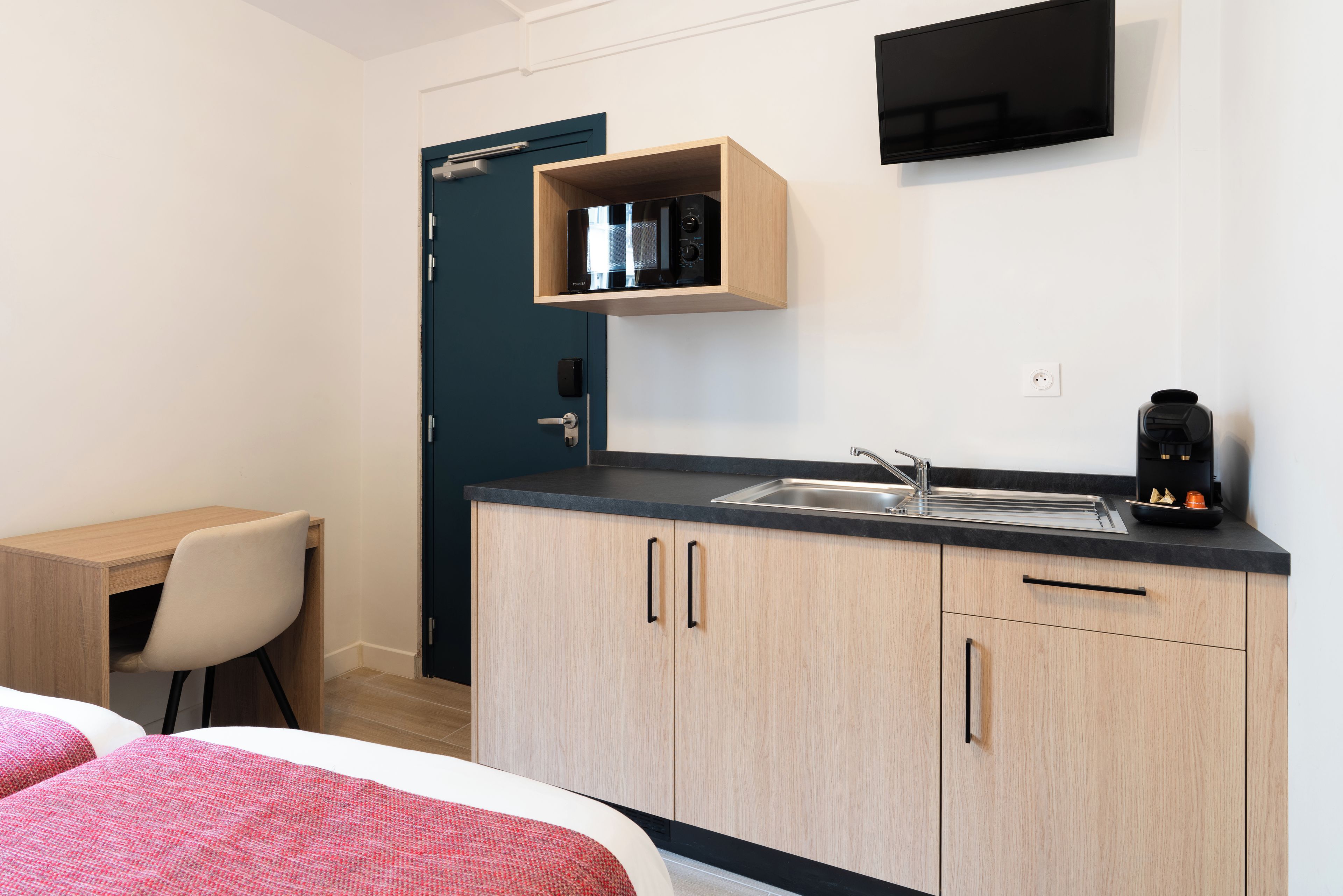 A compact hotel room with a kitchenette, a bed, a desk, and a chair.