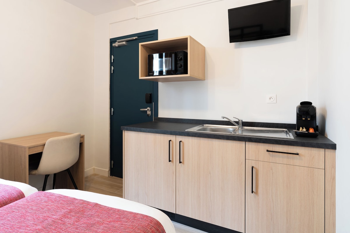 A compact hotel room with a kitchenette, a bed, a desk, and a chair.