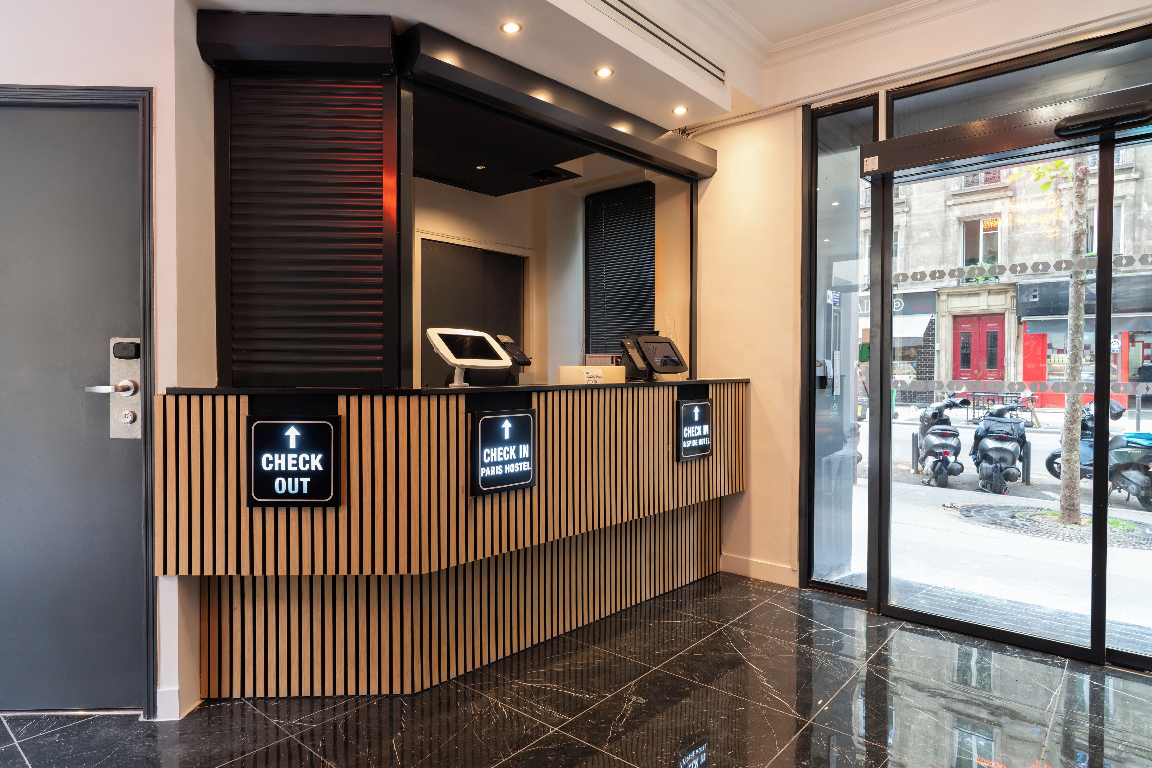 Un comptoir d’accueil avec les panneaux « ENREGISTREMENT » et « DÉPART », un kiosque numérique et une vue sur les scooters garés à l’extérieur.