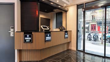 A reception desk with 'CHECK IN' and 'CHECK OUT' signs, a digital kiosk, and a view of parked scooters outside.