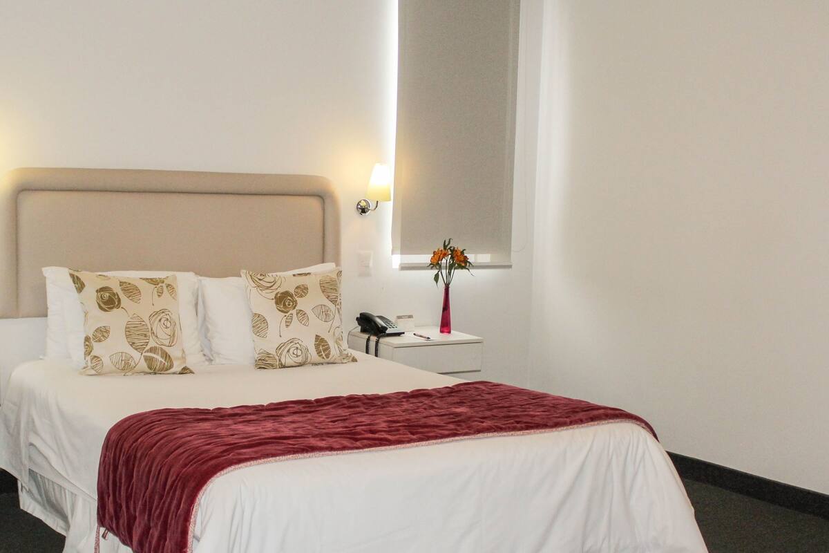 Habitación de hotel con cama, mesitas de noche, una jarra con flores y lámparas de pared.
