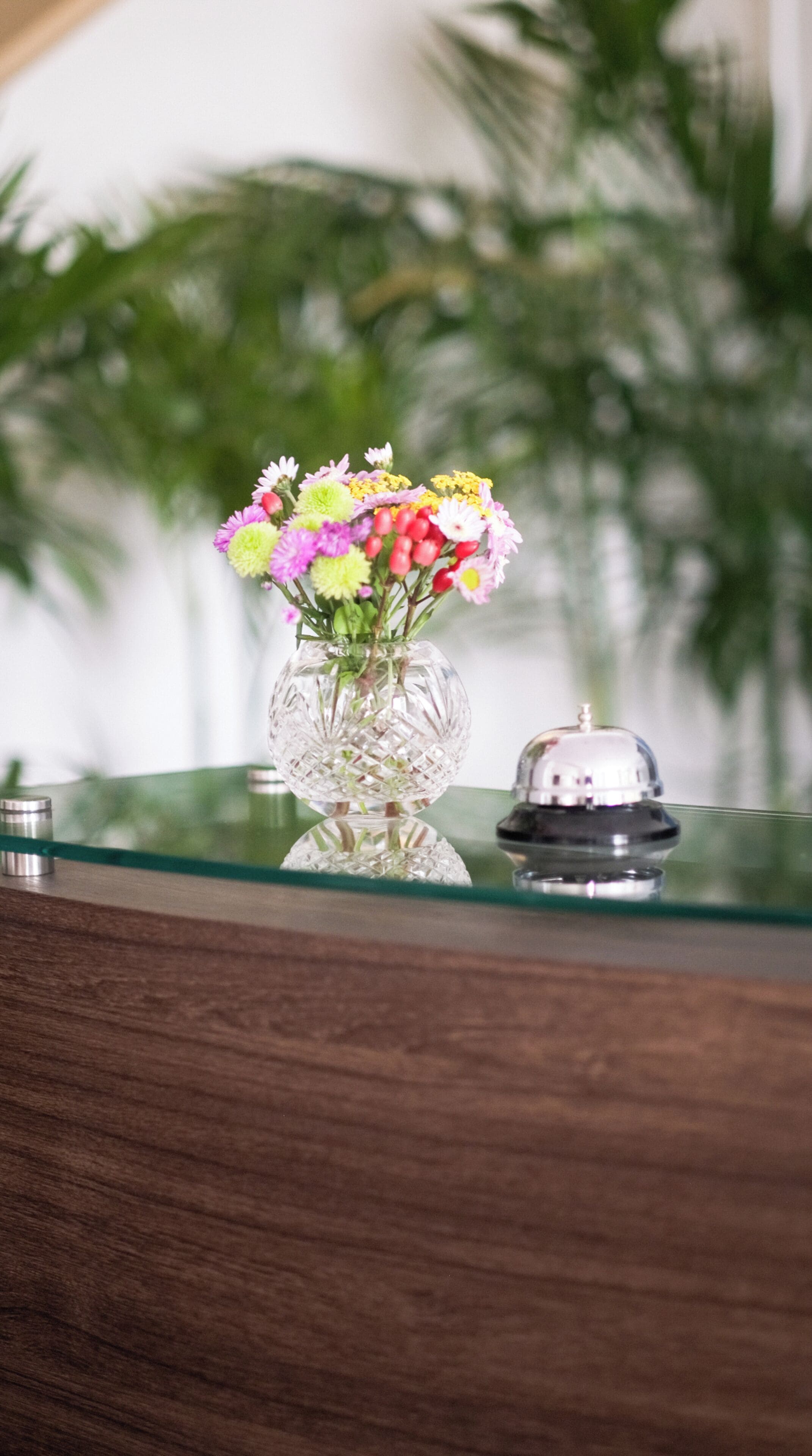 A glass vase with a variety of flowers on a reflective surface, a small bell, and a blurred background with greenery.