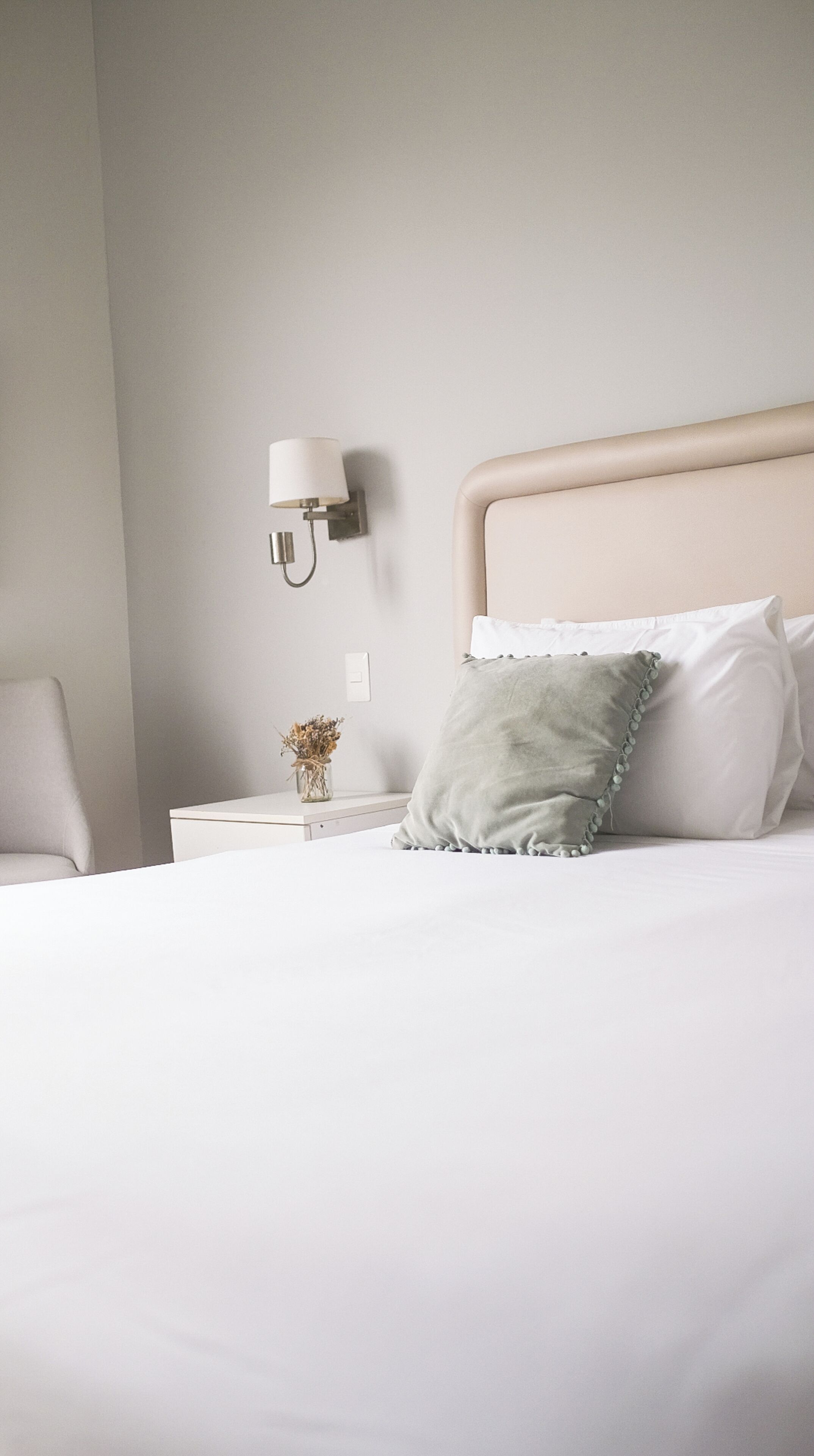 A neatly made bed with a headboard, a bedside lamp, and a small vase with dried flowers on a nightstand.
