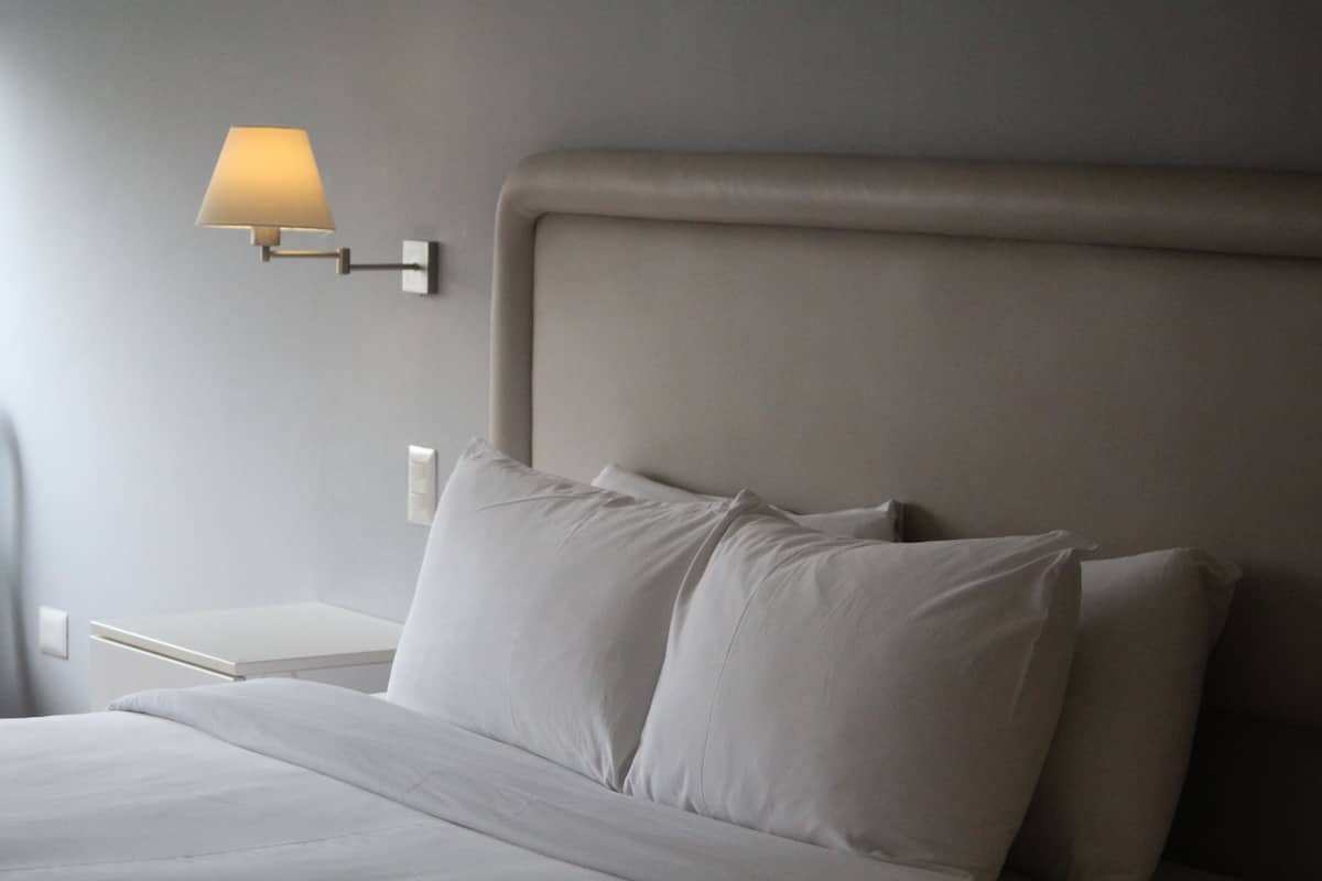 Una cama bien tendida con sábanas y almohadas blancas, una mesita de noche con una lámpara y una lámpara de pared.