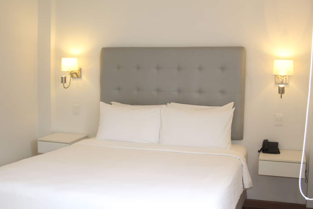 Una cama bien hecha con un cabecero gris, dos mesitas de noche con lámparas, y un teléfono sobre una de ellas.