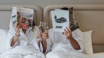 Dos personas vestidas de blanco sentadas en una cama con ropa de cama blanca, sosteniendo revistas.