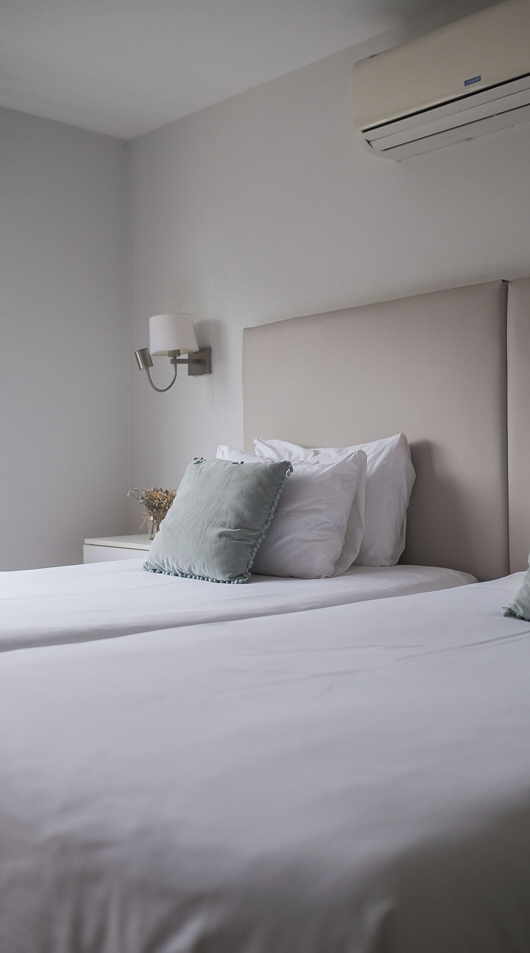 A neatly made bed with white bedding and two pillows, a bedside table with a vase of dried flowers, and a wall-mounted air conditioning unit.