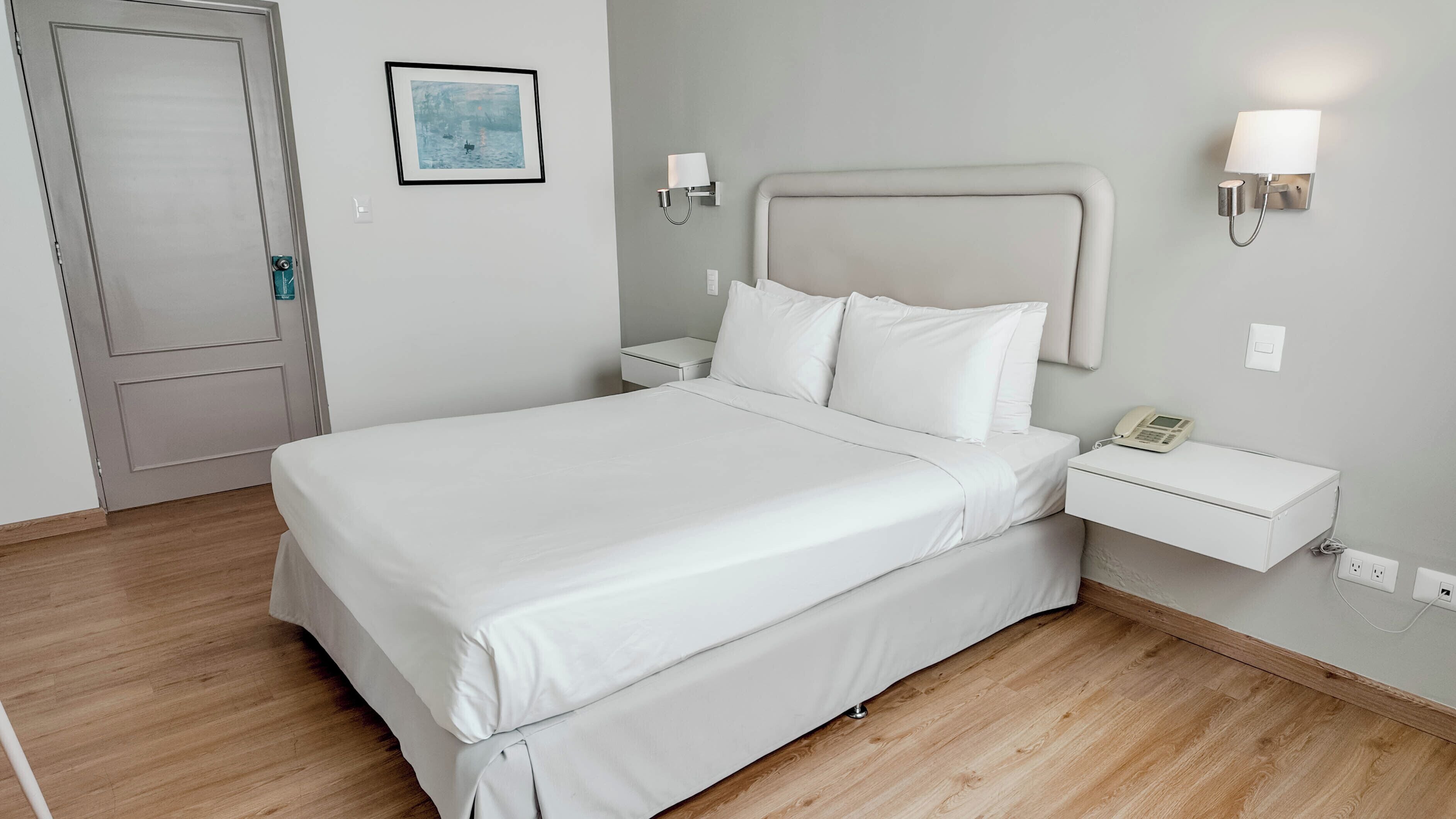 A neatly arranged hotel room with a large bed, a bedside table with a lamp, and a small bedside table with a phone.
