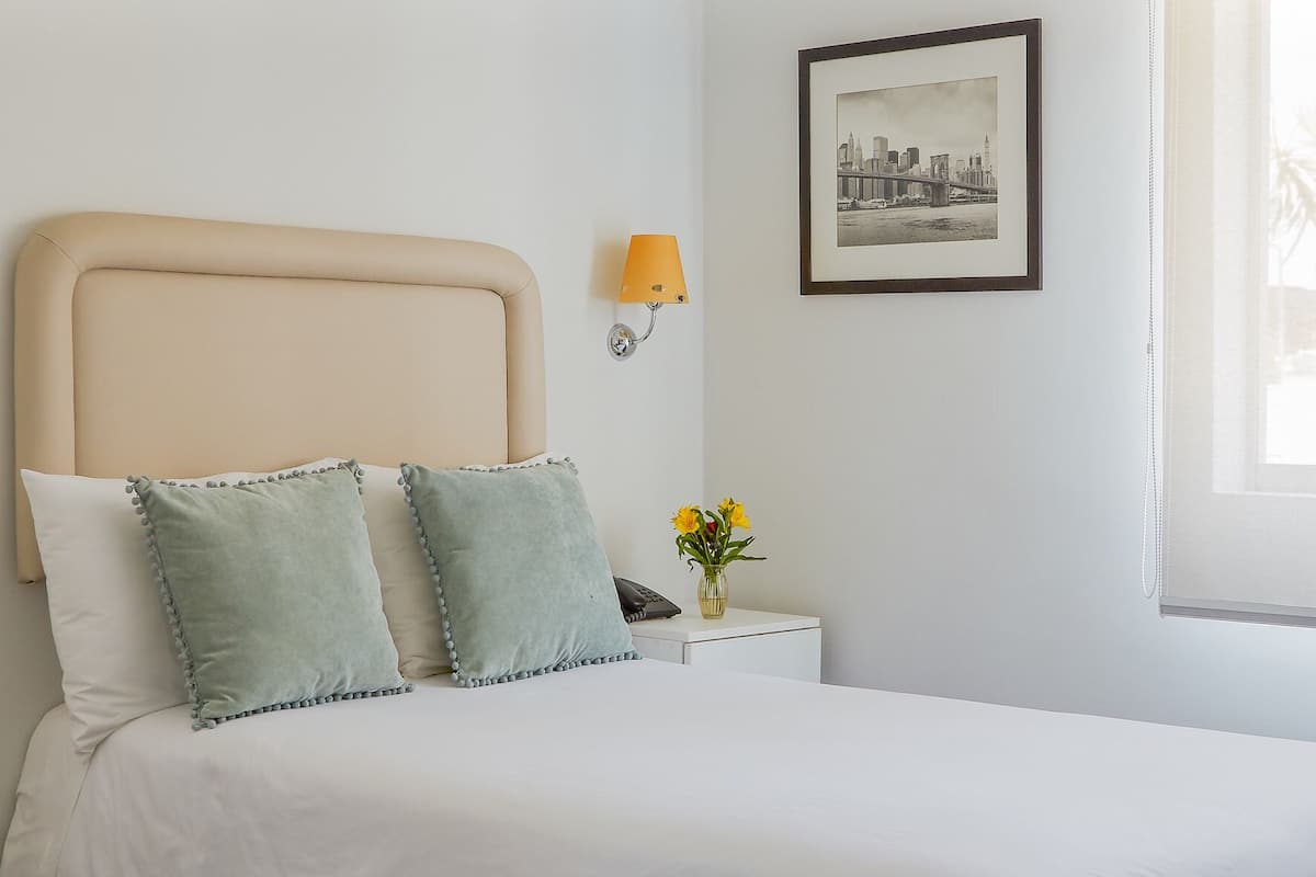 Una cama bien hecha con cabecera, una lámpara de noche, un cuadro en la pared y una ventana con vistas a palmeras.