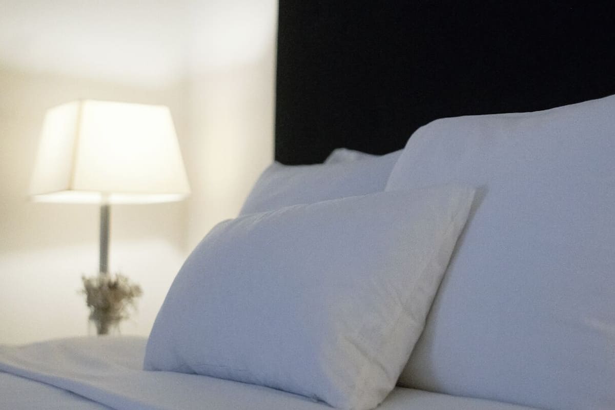 Una cama bien hecha con sábanas y almohadas blancas, un cabecero negro y una lámpara de noche con pantalla blanca.