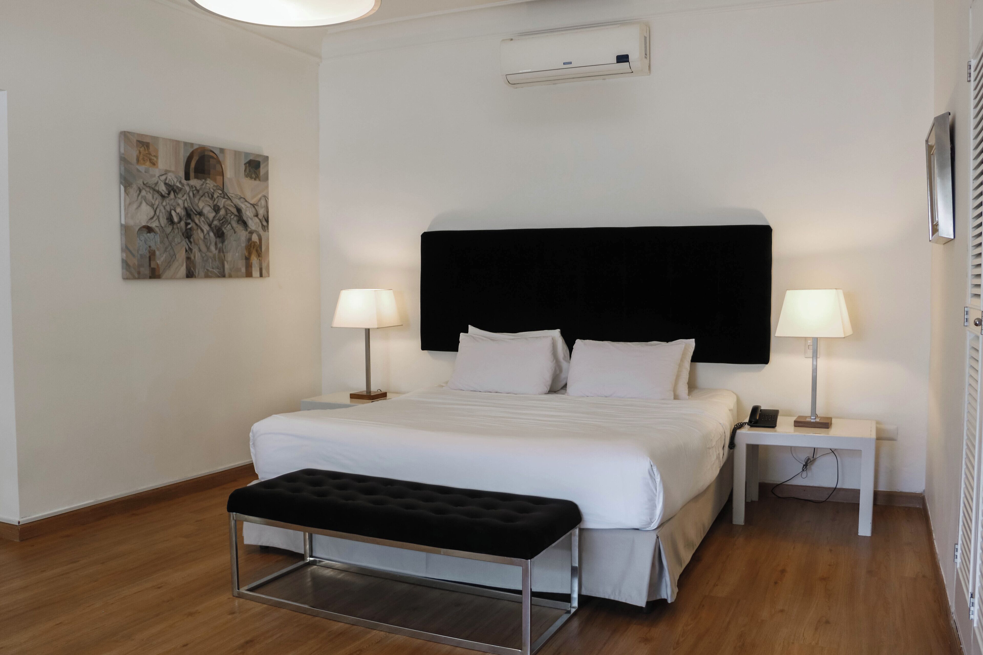 A modern hotel room with a large bed, two bedside tables with lamps, a bench, and a painting on the wall.