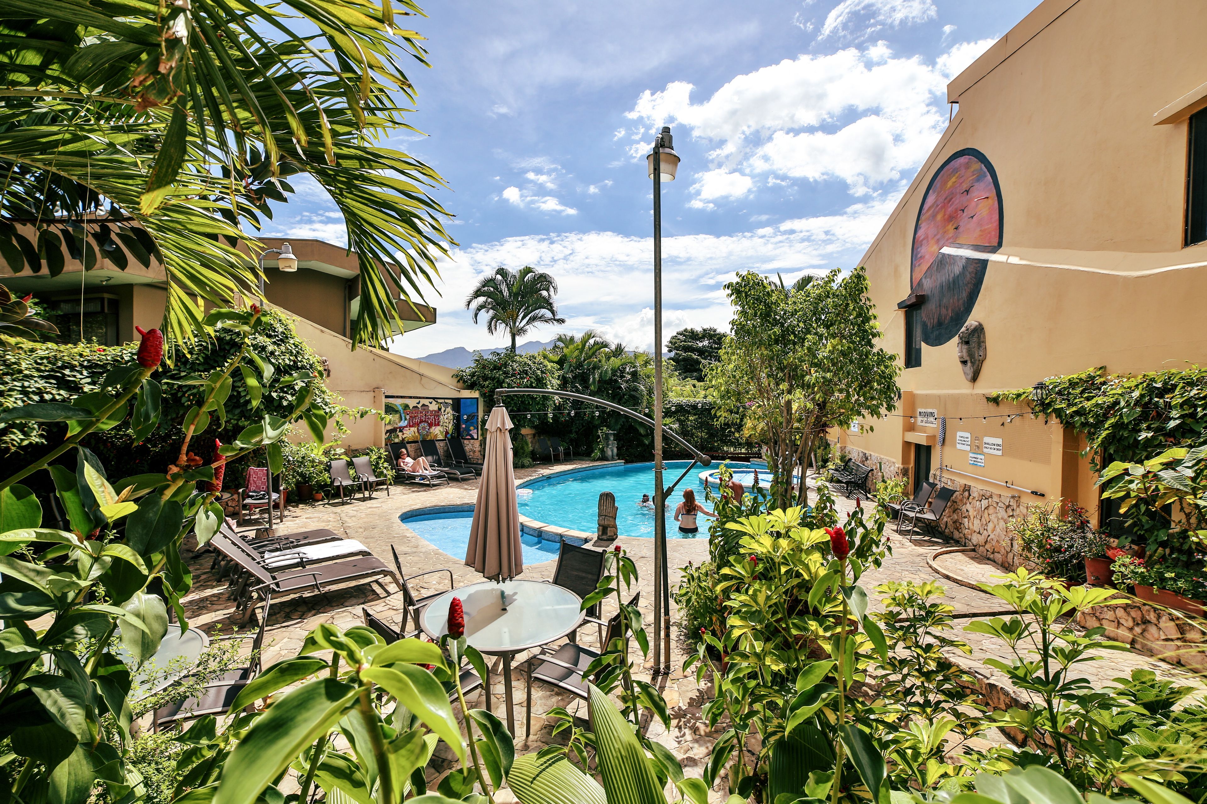 A pool area with lounge chairs and umbrellas, surrounded by greenery and a building with a mural.