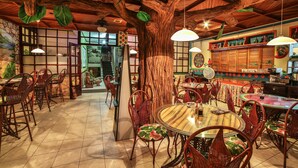 A restaurant with round tables and wicker chairs, a large tree trunk centerpiece, and traditional decor. - Adventure Inn (Ulloa)