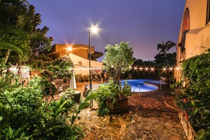 A pool area with a stone pathway, surrounded by lush greenery and a building with a curved roof.