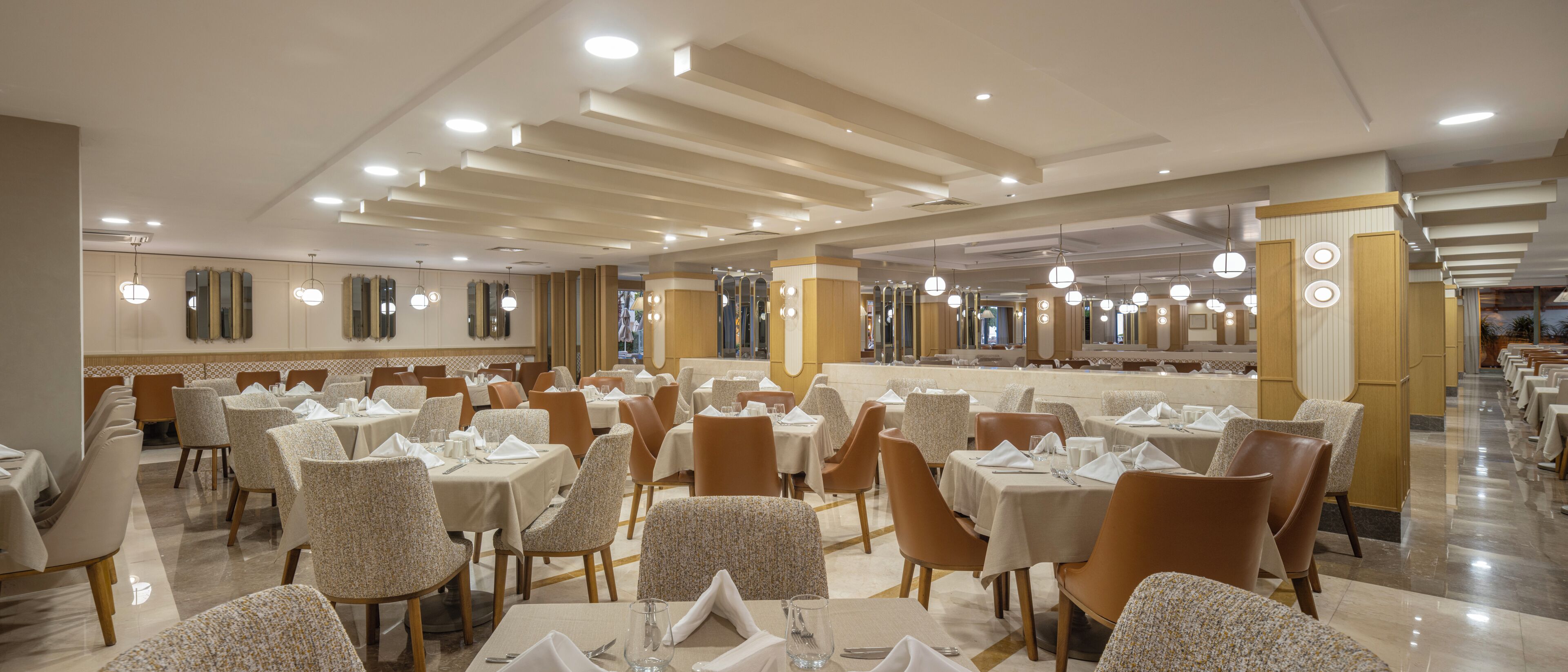 Un amplio salón comedor de hotel con numerosas mesas y sillas, una barra central y un diseño moderno en el techo.
