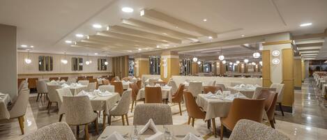 Un amplio salón comedor de hotel con numerosas mesas y sillas, una barra central y un diseño moderno en el techo.