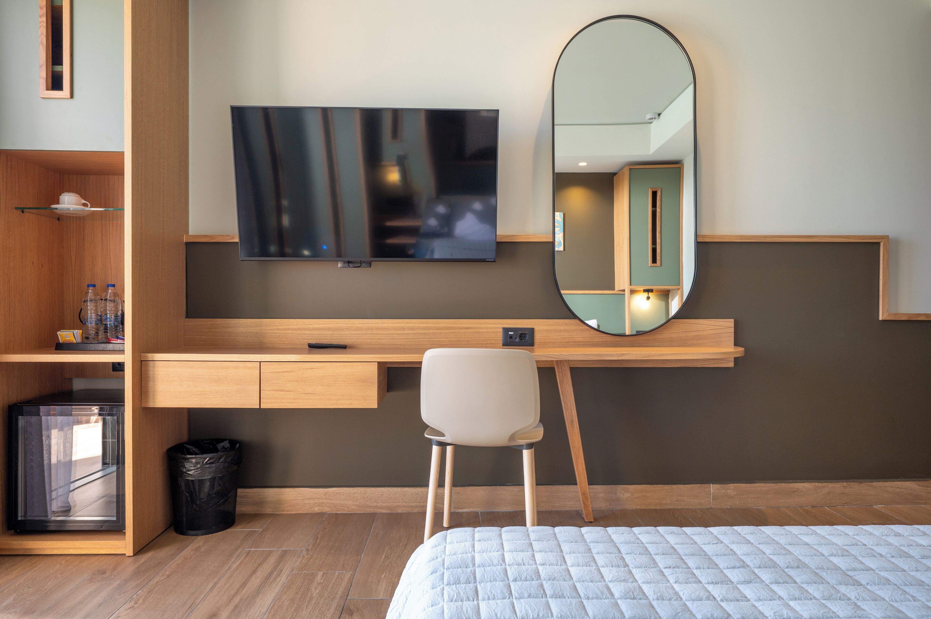 A hotel room with a bed, a desk with a chair, a TV, a mirror, and a trash bin.